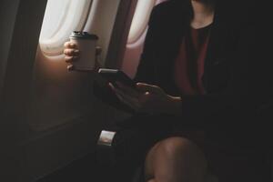 Blonde female tourist checking incoming notification on smartphone sitting on seat of airplane with netbook.Young businesswoman share media from telephone on laptop computer during plane flight photo