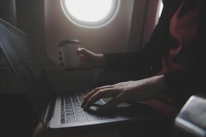 Blonde female tourist checking incoming notification on smartphone sitting on seat of airplane with netbook.Young businesswoman share media from telephone on laptop computer during plane flight photo
