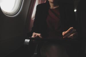 Blonde female tourist checking incoming notification on smartphone sitting on seat of airplane with netbook.Young businesswoman share media from telephone on laptop computer during plane flight photo