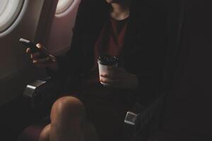 Blonde female tourist checking incoming notification on smartphone sitting on seat of airplane with netbook.Young businesswoman share media from telephone on laptop computer during plane flight photo