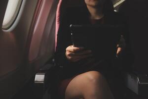 Blonde female tourist checking incoming notification on smartphone sitting on seat of airplane with netbook.Young businesswoman share media from telephone on laptop computer during plane flight photo