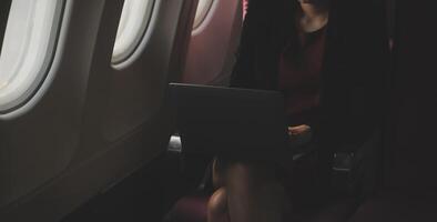 Blonde female tourist checking incoming notification on smartphone sitting on seat of airplane with netbook.Young businesswoman share media from telephone on laptop computer during plane flight photo