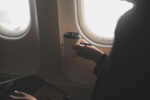 Blonde female tourist checking incoming notification on smartphone sitting on seat of airplane with netbook.Young businesswoman share media from telephone on laptop computer during plane flight photo