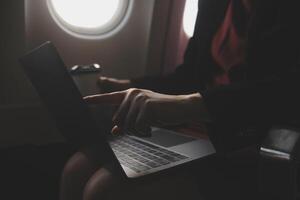 Blonde female tourist checking incoming notification on smartphone sitting on seat of airplane with netbook.Young businesswoman share media from telephone on laptop computer during plane flight photo
