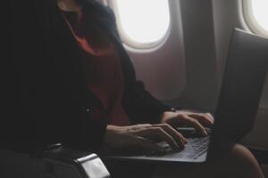 Blonde female tourist checking incoming notification on smartphone sitting on seat of airplane with netbook.Young businesswoman share media from telephone on laptop computer during plane flight photo