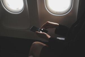 Blonde female tourist checking incoming notification on smartphone sitting on seat of airplane with netbook.Young businesswoman share media from telephone on laptop computer during plane flight photo