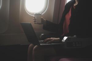 Blonde female tourist checking incoming notification on smartphone sitting on seat of airplane with netbook.Young businesswoman share media from telephone on laptop computer during plane flight photo