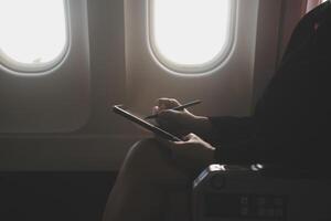 Blonde female tourist checking incoming notification on smartphone sitting on seat of airplane with netbook.Young businesswoman share media from telephone on laptop computer during plane flight photo