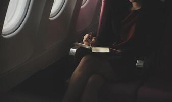 Blonde female tourist checking incoming notification on smartphone sitting on seat of airplane with netbook.Young businesswoman share media from telephone on laptop computer during plane flight photo