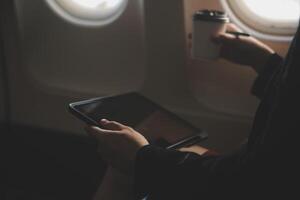 Blonde female tourist checking incoming notification on smartphone sitting on seat of airplane with netbook.Young businesswoman share media from telephone on laptop computer during plane flight photo