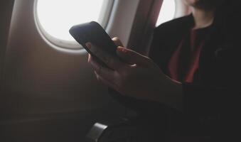 Blonde female tourist checking incoming notification on smartphone sitting on seat of airplane with netbook.Young businesswoman share media from telephone on laptop computer during plane flight photo