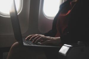Blonde female tourist checking incoming notification on smartphone sitting on seat of airplane with netbook.Young businesswoman share media from telephone on laptop computer during plane flight photo