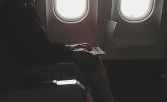 Blonde female tourist checking incoming notification on smartphone sitting on seat of airplane with netbook.Young businesswoman share media from telephone on laptop computer during plane flight photo
