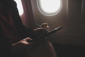 Blonde female tourist checking incoming notification on smartphone sitting on seat of airplane with netbook.Young businesswoman share media from telephone on laptop computer during plane flight photo