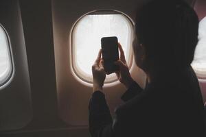Blonde female tourist checking incoming notification on smartphone sitting on seat of airplane with netbook.Young businesswoman share media from telephone on laptop computer during plane flight photo
