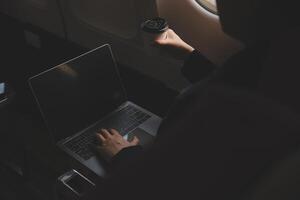 Blonde female tourist checking incoming notification on smartphone sitting on seat of airplane with netbook.Young businesswoman share media from telephone on laptop computer during plane flight photo