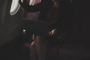 Blonde female tourist checking incoming notification on smartphone sitting on seat of airplane with netbook.Young businesswoman share media from telephone on laptop computer during plane flight photo
