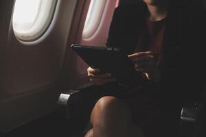Blonde female tourist checking incoming notification on smartphone sitting on seat of airplane with netbook.Young businesswoman share media from telephone on laptop computer during plane flight photo