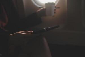 Blonde female tourist checking incoming notification on smartphone sitting on seat of airplane with netbook.Young businesswoman share media from telephone on laptop computer during plane flight photo