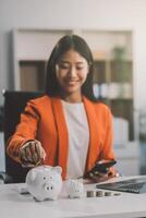 Beautiful Asian businesswoman insert coins in a pin piggy bank on the table. saving money, investment, currency concept photo