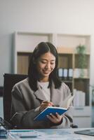 Confident millennial indian female business analyst financial advisor preparing statistic report studying documents on work desk, browsing information online using pc, writing out notes on paper sheet photo