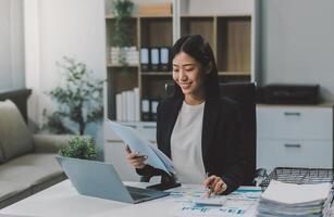Confident millennial indian female business analyst financial advisor preparing statistic report studying documents on work desk, browsing information online using pc, writing out notes on paper sheet photo