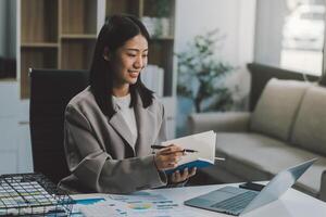 Confident millennial indian female business analyst financial advisor preparing statistic report studying documents on work desk, browsing information online using pc, writing out notes on paper sheet photo