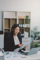 Confident millennial indian female business analyst financial advisor preparing statistic report studying documents on work desk, browsing information online using pc, writing out notes on paper sheet photo