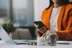 Beautiful Asian businesswoman insert coins in a pin piggy bank on the table. saving money, investment, currency concept photo