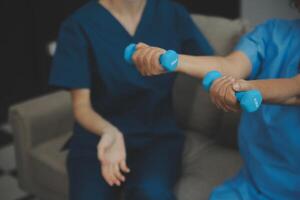 Old woman training with physiotherapist using dumbbells at home. Therapist assisting senior woman with exercises in nursing home. Elderly patient using dumbbells with outstretched arms. photo
