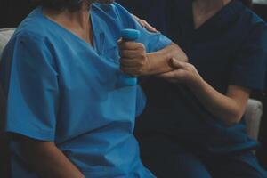 Old woman training with physiotherapist using dumbbells at home. Therapist assisting senior woman with exercises in nursing home. Elderly patient using dumbbells with outstretched arms. photo
