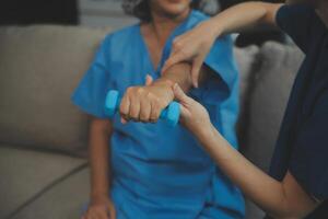 Old woman training with physiotherapist using dumbbells at home. Therapist assisting senior woman with exercises in nursing home. Elderly patient using dumbbells with outstretched arms. photo