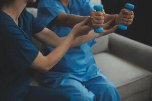 Old woman training with physiotherapist using dumbbells at home. Therapist assisting senior woman with exercises in nursing home. Elderly patient using dumbbells with outstretched arms. photo