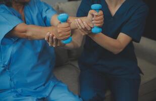 Old woman training with physiotherapist using dumbbells at home. Therapist assisting senior woman with exercises in nursing home. Elderly patient using dumbbells with outstretched arms. photo