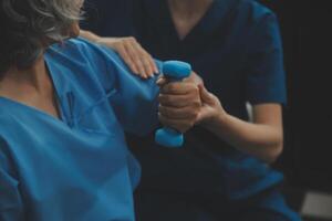 Old woman training with physiotherapist using dumbbells at home. Therapist assisting senior woman with exercises in nursing home. Elderly patient using dumbbells with outstretched arms. photo