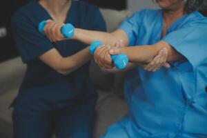 Old woman training with physiotherapist using dumbbells at home. Therapist assisting senior woman with exercises in nursing home. Elderly patient using dumbbells with outstretched arms. photo
