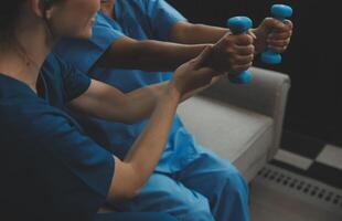 Old woman training with physiotherapist using dumbbells at home. Therapist assisting senior woman with exercises in nursing home. Elderly patient using dumbbells with outstretched arms. photo