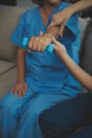 Old woman training with physiotherapist using dumbbells at home. Therapist assisting senior woman with exercises in nursing home. Elderly patient using dumbbells with outstretched arms. photo
