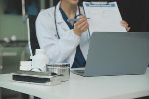 A beautiful and professional Asian female doctor is working on her medical cases on her laptop or making a call medical consultation with a patient while sitting in her office. photo