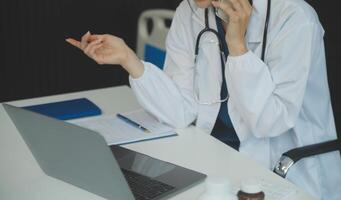 A beautiful and professional Asian female doctor is working on her medical cases on her laptop or making a call medical consultation with a patient while sitting in her office. photo