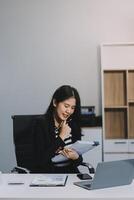 Charming Young asian businesswoman sitting on laptop computer in the office, making report calculating balance Internal Revenue Service checking document. photo