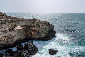 The ocean is calm and the sky is cloudy. The water is a deep blue color. The rocks are jagged and the waves are small. photo