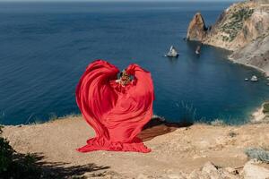 A woman in a red dress is standing on a rocky beach, with the ocean in the background. The woman is wearing a red dress that is flowing out in a heart shape, giving the impression of a romantic photo