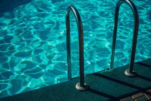A metal ladder is in the water. The water is clear and calm. The ladder is in the middle of the pool. photo