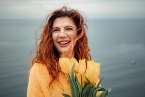 Portrait of a happy woman with hair flying in the wind against the backdrop of mountains and sea. Holding a bouquet of yellow tulips in her hands, wearing a yellow sweater photo