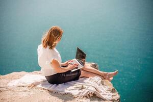 Freelance woman working on a laptop by the sea, typing away on the keyboard while enjoying the beautiful view, highlighting the idea of remote work. photo