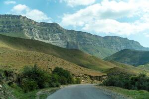 A road runs through a valley with mountains in the background. The road is surrounded by trees and grass, and the sky is clear and blue. photo