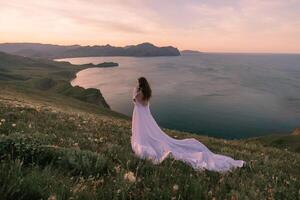 A woman in a white dress stands on a hill overlooking the ocean photo