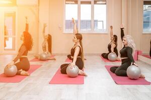A group of six athletic women doing pilates or yoga on pink mats in front of a window in a beige loft studio interior. Teamwork, good mood and healthy lifestyle concept. photo