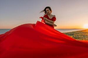 woman red dress standing grassy hillside. The sun is setting in the background, casting a warm glow over the scene. The woman is enjoying the beautiful view and the peaceful atmosphere. photo